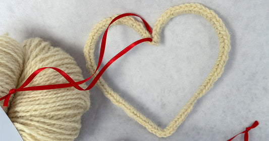 Gehäkeltes Deko-Herz aus naturfarbener Wolle mit rotem Satinband, daneben ein Knäuel Wolle – DIY-Idee als liebevolle Dekoration oder Geschenk.